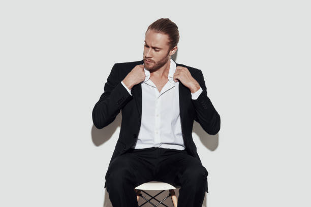 istock Perfect style. Handsome young man in full suit looking away and adjusting jacket while sitting against grey background 1157120006