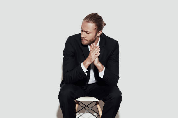 istock Taking time to think. Handsome young man in full suit looking away and keeping hands clasped while sitting against grey background 1157121189