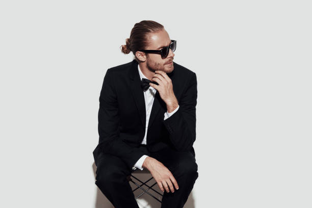 istock Thinking about... Charming young man in full suit keeping hand on chin and looking away while sitting against grey background 1157121214