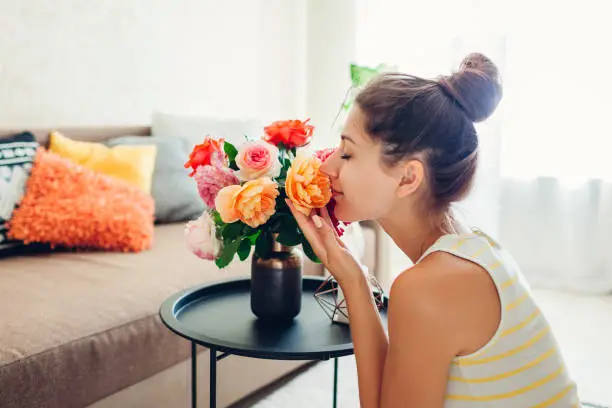 Photo of Woman smelling fresh roses in vase on table. Housewife taking care of coziness in apartment. Interior and decor