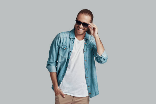 istock Simply charming. Handsome young man adjusting sunglasses and smiling while standing against grey background 1157130022
