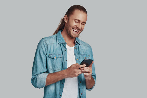 istock Social gathering. Handsome young man using smart phone and smiling while standing against grey background 1157130885