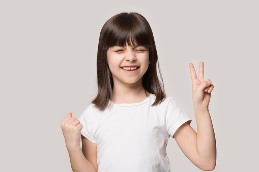 On beige studio background pose little girl in white tshirt got what she wanted feel excitement show V victory symbol, Yes gesture, demonstrating give peace sign, dream came true, celebration concept