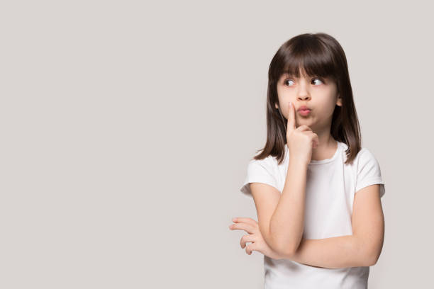 Thoughtful little girl looking away thinking isolated on grey background Thoughtful little girl six years old daughter look away on copy space thinking isolated on grey beige background, funny kid pout lips hold finger near mouth conceived some kind of prank, concept image unstable face expression from little cute kid stock pictures, royalty-free photos & images