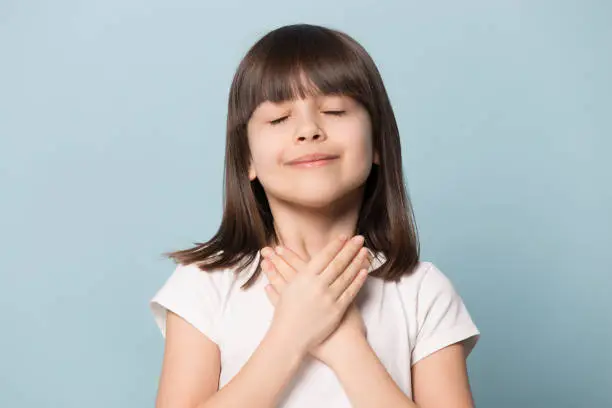 Head shot appreciative little brown-haired girl closed eyes holding hands on chest symbol of deep grateful feelings isolated on blue background, adopted child new parents, Gratitude Day concept image