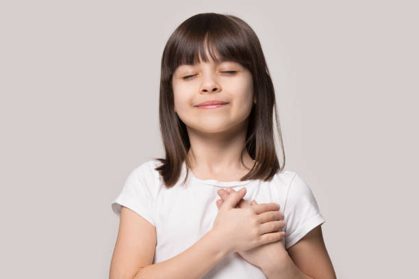 Little girl closed eyes hold hand on chest feels gratitude Little sincere adorable girl closed eyes holding hands on chest feeling gratitude pose isolated on sandy color beige background, arms on heart gesture of love appreciation gratitude, adoption concept worshipper photos stock pictures, royalty-free photos & images