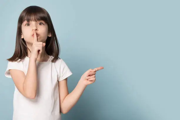 Adorable six years old little girl holding finger on lips symbol of hush gesture of asking to be quiet. Silence or secret concept image isolated on blue studio background with copy free space for text