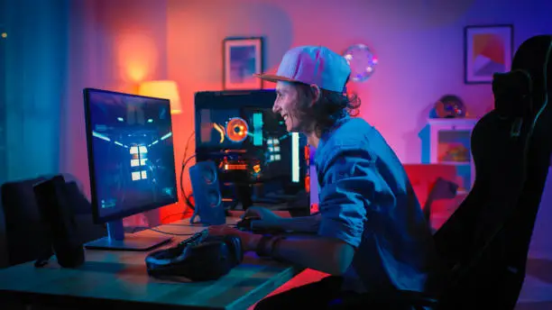 Professional Gamer Playing First-Person Shooter Online Video Game on His Powerful Personal Computer with Colorful Neon Led Lights. Young Man is Wearing a Cap. Living Room Lit with Warm Red Lamps. Evening.