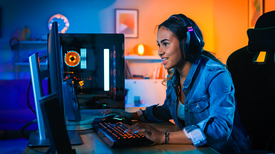 Pretty and Excited Black Gamer Girl en los auriculares está jugando en primera persona Shooter videojuego en línea en su ordenador. La habitación y el PC tienen luces led de neón de colores. Noche acogedora en casa. photo