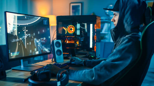 professional gamer and streamer playing first-person shooter online video game on his cool personal computer. young man is wearing a cap and hood. room and pc have colorful neon led lights. cozy evening at home. - people joy relaxation concentration imagens e fotografias de stock