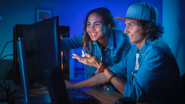 istock Two Blogger Friends or a Couple Actively Discuss Something on a Computer Screen. Pretty Black Girl Points to the Screen and Young Man Agrees with her Creative Ideas. Cozy Room is Lit with Blue Neon Light. 1157159218
