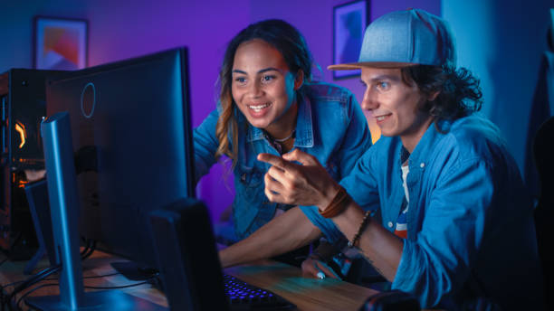 Pretty Black Girl Actively Talks and Explains Something to Her Friend and Points on a Computer Screen. They Have a Discussion about New Creative Ideas. Cozy Room is Lit with Warm and Neon Light.