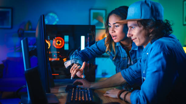 istock Two Blogger Friends or a Couple Actively Discuss Something on a Computer Screen. Pretty Black Girl Points to the Screen and Young Man Agrees with her Creative Ideas. Cozy Room is Lit with Green Neon Light. 1157159299