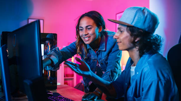istock Two Blogger Friends or a Couple Actively Discuss Something on a Computer Screen. Pretty Black Girl Points to the Screen and Young Man Agrees with her Creative Ideas. Cozy Room is Lit with Ping Neon Light. 1157159372