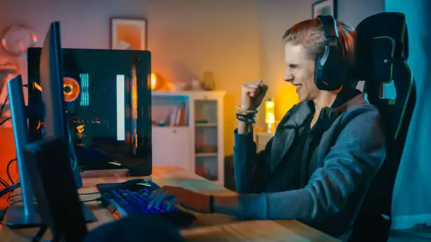 Excited Gamer Playing and Winning in First-Person Shooter Online Video Game on His Personal Computer. Room and PC have Colorful Neon Led Lights. Young Man is Wearing Earphones. Cozy Evening at Home.