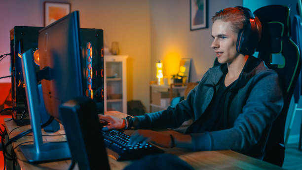 istock Happy Gamer in Headset with a Mic Playing Shooter Online Video Game on His Personal Computer. Room and PC have Colorful Neon Led Lights. Cozy Evening at Home. 1157160129