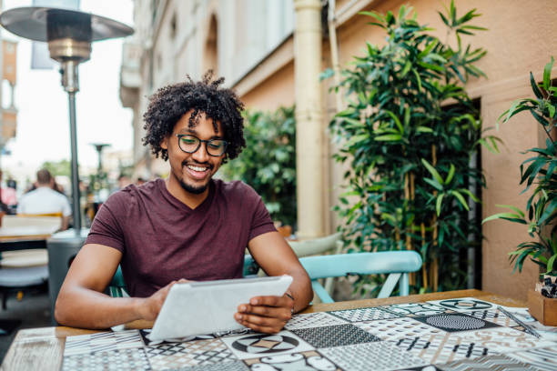 Afro American man using tablet in coffee shop Smiling African student texting outside, sitting in coffee shop cafe tablet stock pictures, royalty-free photos & images