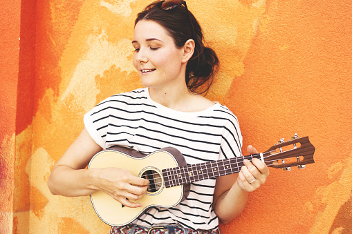 Portrait of a singing brunette hipster girl in a striped T-shirt with a guitar ukulele against the background of the urban orange wall, summer vacation concept. Copy space for text
