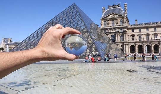Paris, France - August 18, 2018: Pyramid of Louvre Museum and a big glass sphere on the hand of a man