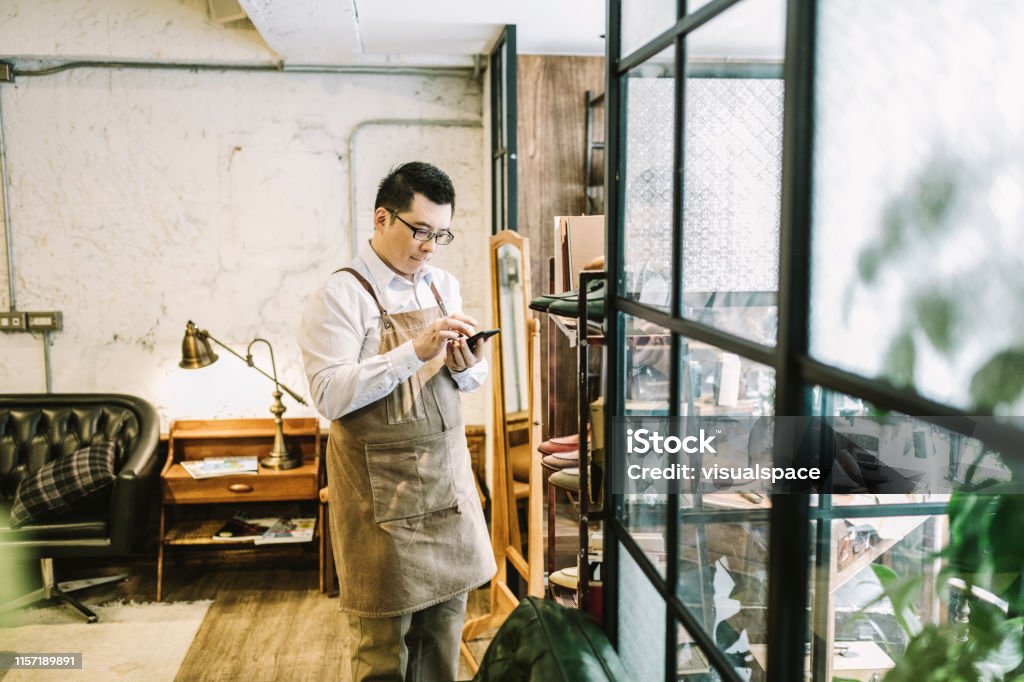Owner answering call while standing by window Smll business owner sending voice message while standing by window. Entrepreneur using smart phone while looking away. He wearing apron in store. Small Business Stock Photo