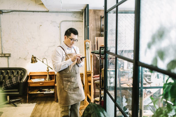 Owner answering call while standing by window Smll business owner sending voice message while standing by window. Entrepreneur using smart phone while looking away. He wearing apron in store. taipei photos stock pictures, royalty-free photos & images