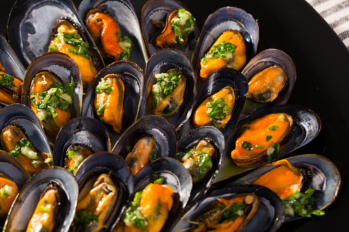Closeup of appetizing steamed mussels with sauce of oil, garlic and parsley on black ceramic dish