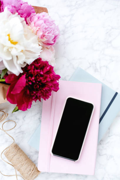 Bouquet of pink peonies, a stack of notebooks and mobile phone on marble stone table with copy space. View from the top. Empty screen for text.