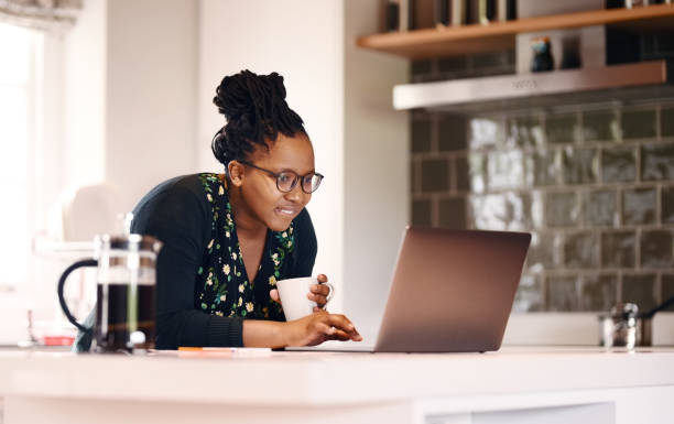 Trying to search some information Shot of a young african american woman with a coffee cup using laptop in kitchen at home searching stock pictures, royalty-free photos & images