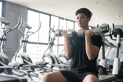 close up young asian man holding dumbbell in hand for playing shoulder press to gaining muscle at gym in the afternoon , healthy lifestyle concept