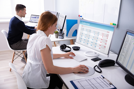 Businesswoman Looking At Calendar On Desktop Computer With Colleague Behind In Office
