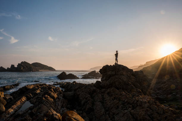 istock Man standing on rocky coastline at beach at sunset 1157270813