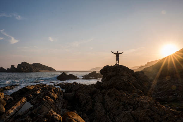 Distant image of man with arms outstretched standing on rock. Male is enjoying the view of Plettenberg Bay against sky during sunset. He is on summer vacation at beach.
