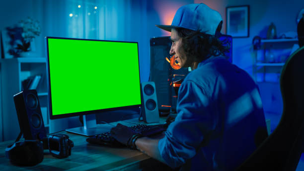 Professional Gamer Playing Online Video Game on His Powerful Personal Computer with Colorful Neon Led Lights. Green Screen Mock Up. Young Man is Wearing a Cap. Living Room with Warm Lamps. Evening. Professional Gamer Playing Online Video Game on His Powerful Personal Computer with Colorful Neon Led Lights. Green Screen Mock Up. Young Man is Wearing a Cap. Living Room with Warm Lamps. Evening. chroma key photos stock pictures, royalty-free photos & images
