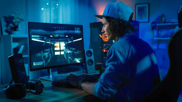 Professional Gamer Playing First-Person Shooter Online Video Game on His Powerful Personal Computer with Colorful Neon Led Lights. Young Man is Wearing a Cap. Living Room Lit in Low Key Style. Evening.