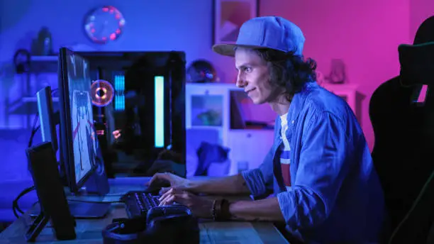 Professional Gamer Playing First-Person Shooter Online Video Game on His Powerful Personal Computer with Colorful Neon Led Lights. Young Man is Wearing a Cap. Living Room Lit with Pink Neom Light.
