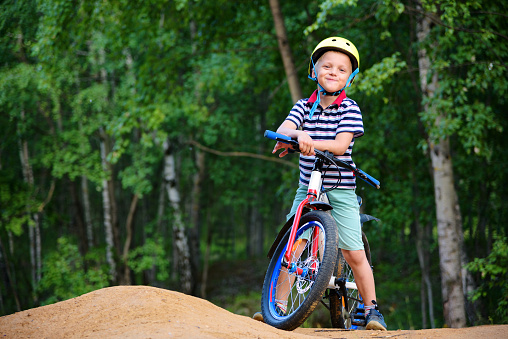 Boy cyclist standing in full growth near his bike against the background of the forest. Wearing a striped polo, blue shorts and a yellow helmet.