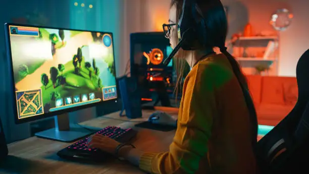 Photo of Excited and Concentrated Gamer Girl in Glasses and Headset with a Mic Playing Online Strategy Video Game on Her Personal Computer. Room and PC have Colorful Warm Neon Led Lights. Cozy Evening at Home.