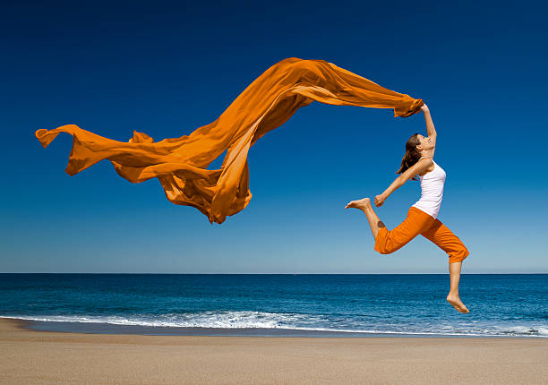 Woman with orange scarf jumping in the beach A woman wearing orange pants and a white top is on a beach.  The water is blue.  The woman jumps into the air, and she kicks her right leg behind her.  She has an orange scarf in her left hand, and it floats above and behind her against a blue sky. people jumping sea beach stock pictures, royalty-free photos & images