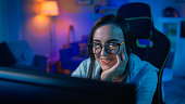 Beautiful Happy and Sentimental Young Girl Blogger Watching Videos on a Computer. She Has Dark Hair and Wears Glasses. Screen Adds Reflections to Her Face. Cozy Room is Lit with Warm Light.