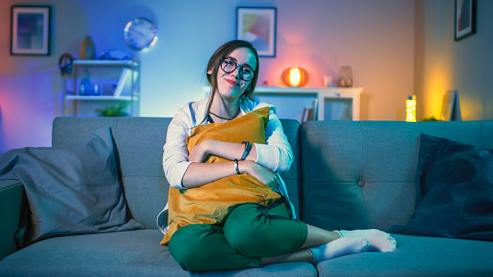 Beautiful Emotional Young Girl in Glasses Sitting on a Couch and Watching TV at Home. She is Hugging a Pillow. Screen Adds Reflections to Her Face. Cozy Room is Lit with Warm Light.