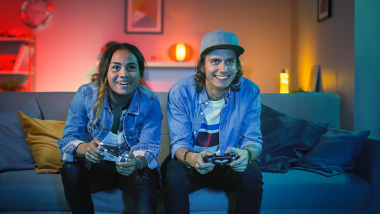 Emocionada Black Gamer Girl y Young Man Sitting on a Couch and Playing Video Games on Console. Juegan con controladores inalámbricos. Habitación acogedora con luz cálida y de neón. photo