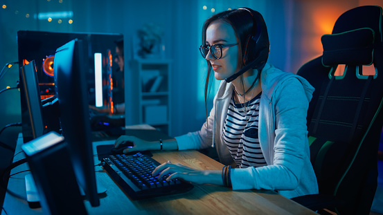 Chica jugadora emocionada en auriculares con un micrófono jugando videojuego en línea en su ordenador personal. Habla con otros jugadores. La habitación y el PC tienen luces led de neón cálidos coloridos. Noche acogedora en casa. photo
