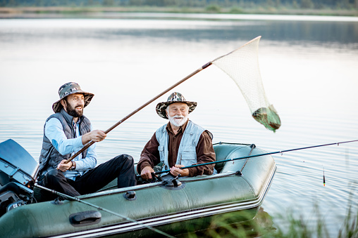 Grandfather with adult son fishing on the inflatable boat, catching fish with net on the lake early in the morning