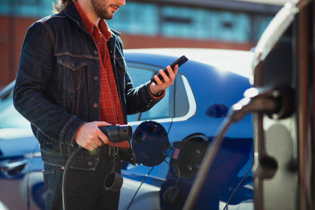 Paying for Electricity With Phone Man plugging in a charger into his electric car while holding his phone, looking down at it, using it to pay for alternative fuel. hybrid car photos stock pictures, royalty-free photos & images