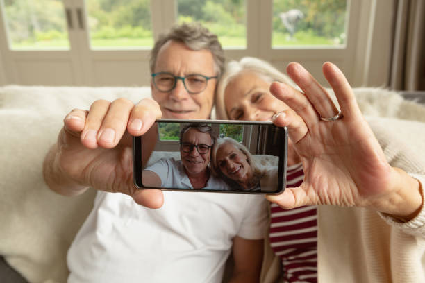 Active senior couple sitting on sofa and taking selfie in a comfortable home Front view of happy active senior Caucasian couple sitting on sofa and taking selfie in a comfortable home photo messaging stock pictures, royalty-free photos & images