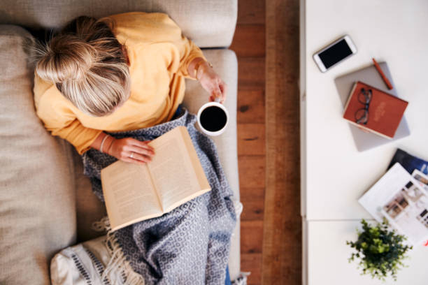 au-dessus de la tête de projectile regardant vers le bas sur la femme à la maison se trouvant sur le livre de lecture et buvant le café - lire photos et images de collection