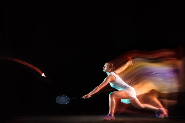 Young woman playing badminton isolated on black background in mixed light. Female model with the racket in action, motion in game with the fire shadows. Concept of sport, movement, healthy lifestyle.