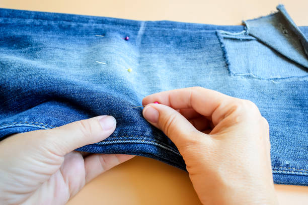 istock Woman hands pin folded in half eans before cutting. Jeans with a large hole on a pant leg below the knee. Making blue denim shorts. 1157323150