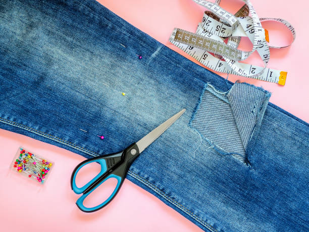 istock Folded in a half blue jeans with large hole on pant leg below the knee, multi coloured headed sewing pins, white tailor tape with centimeters and inches and scissors. 1157323154