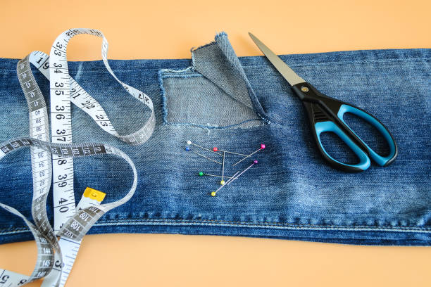 istock Blue jeans with a large hole on a pant leg below the knee, multi coloured headed sewing pins, white tailor tape with inches and centimeters and scissors. 1157323217
